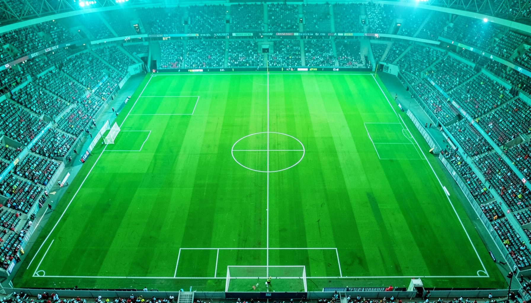A vibrant green football pitch seen from above, with bright white field markings and a turquoise-tinged stadium atmosphere, conveying competitive energy and sportsmanship.